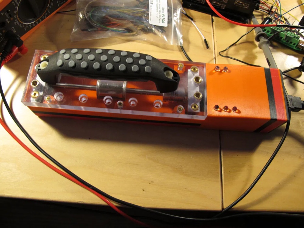 The assembled handheld driver unit: a 3D-printed orange enclosure with an acrylic top plate, gripped handle, and the lead screw and rod visible through the window.