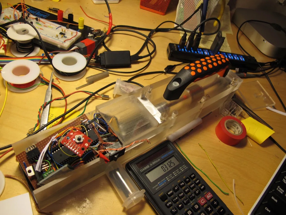 The driver unit opened up on a workbench, showing hand-soldered perfboard with the ATmega microcontroller, wiring, and the motor and lead screw inside a clear acrylic housing.